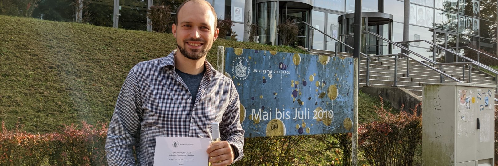 Jan Kopetz steht mit seiner Urkunde des Lehrpreises vor dem Audimax der Universität zu Lübeck.