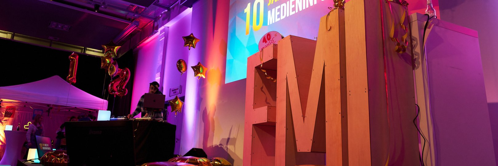 Das Bild zeigt eine bunt beleuchtete Bühne mit den großen Buchstaben E M I darauf stehend, einem DJ-Pult im Hintergrund, goldenen Ballons und eine Leinwand auf der bunt der Schriftzug "10 Jahre Meet-Up der Medieninformatik" abgebildet ist.
