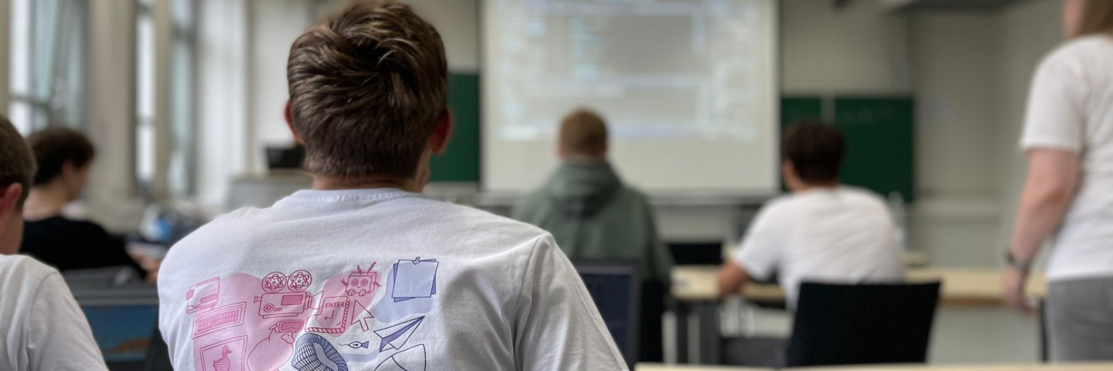 Junge sitzt am Laptop und schaut nach vorne auf Beamer, auf dem Programmcode angezeigt wird.